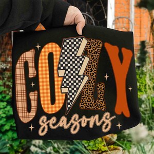 Könnte beinhalten: Schwarzes T-Shirt mit den Worten "COZY seasons" in einer Mischung aus Karomuster, Leopardenmuster und orangefarbenen Buchstaben. Der Blitz ist kariert. Das T-Shirt wird von einer Person gehalten.