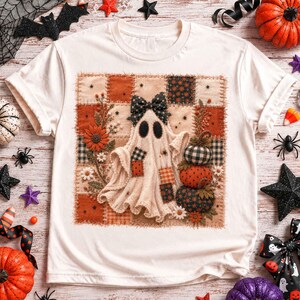 May include: A cream-coloured t-shirt featuring a central square graphic of a patchwork-style ghost wearing a black polka-dot bow. The ghost is surrounded by embroidered-style sunflowers, daisies, and a stack of three patterned pumpkins.