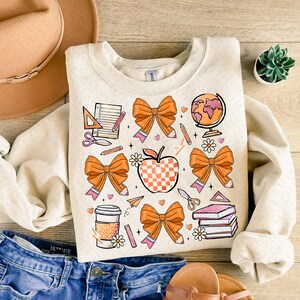 May include: A beige sweatshirt with a colorful back-to-school design. The design features orange bows, a checkered apple, pencils, a globe, a ruler, scissors, a notebook, a coffee cup, a book stack, and a paper airplane.