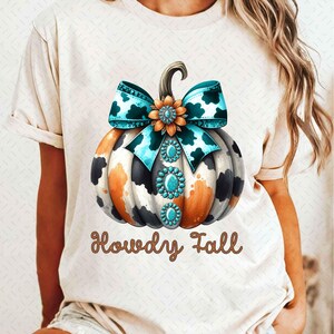 May include: A white t-shirt with a brown, black, and orange cow print pumpkin with a turquoise and white bow and turquoise jewels. The text "Howdy Fall" is printed in brown glitter below the pumpkin.