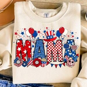 May include: A cream-colored sweatshirt with a red, white, and blue graphic design that says "MAMA" with stars, fireworks, and other patriotic imagery.