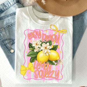 Op de afbeelding: Wit T-shirt met een roze en wit grafisch ontwerp met citroenen, bloemen en de tekst "easy peasy lemon squeezy".