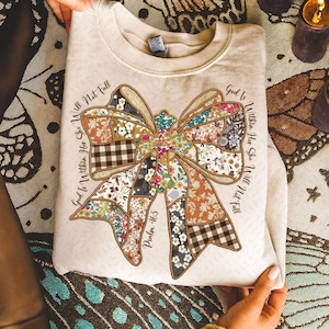 May include: Cream-colored sweatshirt featuring a large bow design composed of various floral and patterned fabrics. The text "God Is Within Her She Will Not Fall" and "Psalm 46:5" are printed around the bow. A cozy, casual top.
