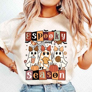 May include: A white t-shirt with a graphic design featuring three ghosts with bows, pumpkins, and the text "E Spooky and Season" in a retro style.