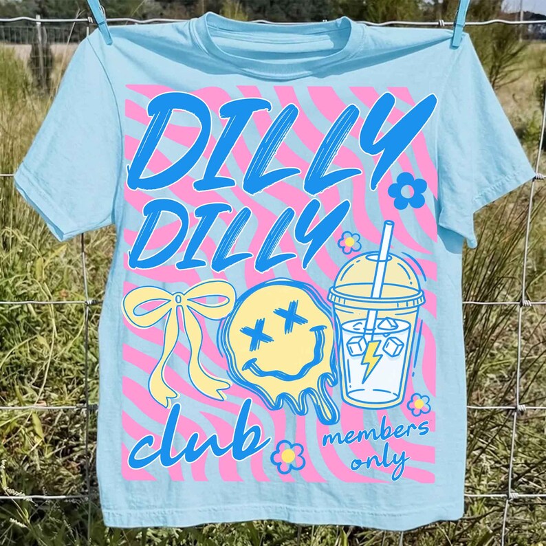 May include: Light blue t-shirt with a colorful graphic. The design includes the words "DILLY DILLY CLUB" in blue and pink, along with a yellow smiley face, a bow, a drink, and the words "members only". The shirt is hanging on a clothesline.