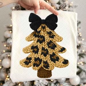 May include: A white fabric with a knitted Christmas tree design. The tree features a black bow at the top, with a pattern of tan and black knit. The tree trunk is brown. The fabric is held up by a hand.