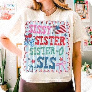 May include: Cream-colored t-shirt with a patriotic design. The design features the words "Sissy Sister Sister-O Sis" in various colors, along with an American flag, stars, and bows. The shirt has a relaxed fit.