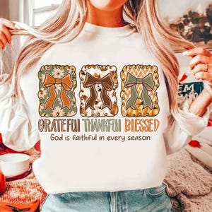 May include: White long-sleeve shirt with three decorative bow designs in fall colors. The bows are in rectangular frames with floral, leaf, and plaid patterns. The text reads "Grateful Thankful Blessed" and "God is faithful in every season."