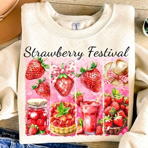 Op de afbeelding: Een cr&egrave;mekleurig sweatshirt met de tekst "Strawberry Festival" in zwart gedrukt. Het ontwerp bevat verschillende illustraties met een aardbeien-thema, waaronder aardbeien, een pot aardbeienjam, een aardbeientaart, een aardbeien-smoothie en een mand met aardbeien.