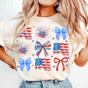 Puede incluir: Una camiseta blanca con un diseño patriótico con estrellas, fuegos artificiales y lazos rojos, blancos y azules. El diseño es de estilo acuarela.
