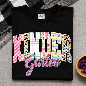 K&ouml;nnte beinhalten: Schwarzes T-Shirt mit den Worten "KINDER Garten" in einem farbenfrohen, verspielten Design. Das Wort "KINDER" hat ein rosa Vichy-Muster, Blumen und Smiley-Akzente. Das Wort "Garten" ist in rosa Schreibschrift.