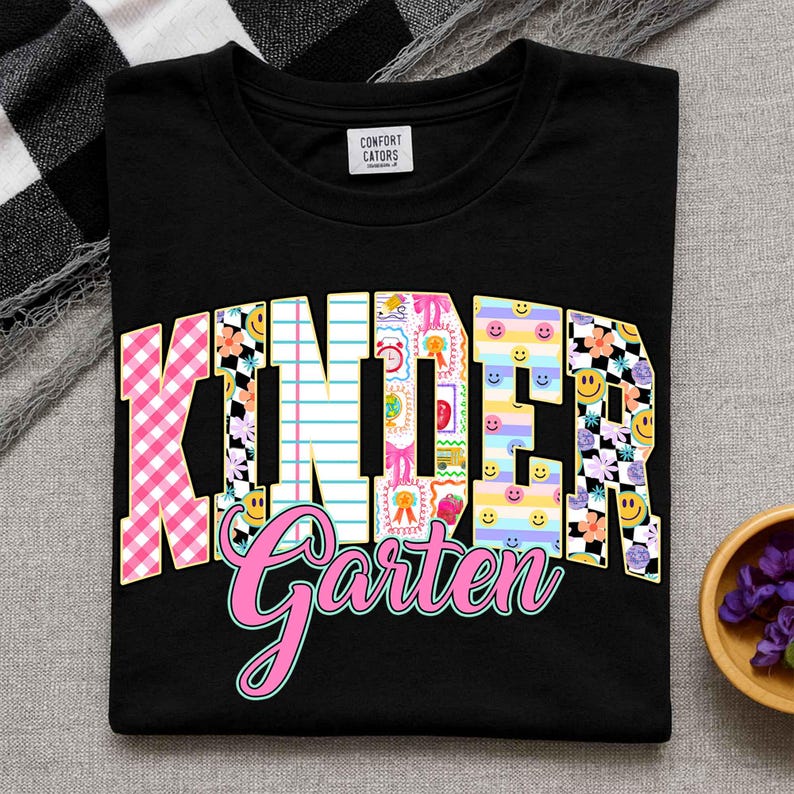 K&ouml;nnte beinhalten: Schwarzes T-Shirt mit den Worten "KINDER Garten" in einem farbenfrohen, verspielten Design. Das Wort "KINDER" hat ein rosa Vichy-Muster, Blumen und Smiley-Akzente. Das Wort "Garten" ist in rosa Schreibschrift.