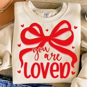 May include: A beige sweatshirt with a red bow and the text "you are loved" in red and pink lettering.