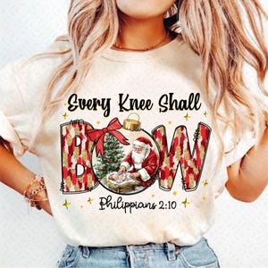 May include: A white t-shirt with a Christmas ornament design. The ornament features a red bow, a green Christmas tree, and Santa Claus holding a baby Jesus. The text "Every Knee Shall Bow" and "Philippians 2:10" are printed on the shirt.