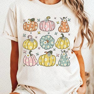 May include: A white t-shirt with a pattern of colorful pumpkins with floral designs. The pumpkins are in various shades of pink, yellow, green, and orange.