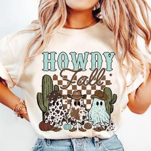 May include: White t-shirt featuring a graphic design of three ghosts wearing cowboy hats and boots. The ghosts are in front of a chequered background with the text "Howdy Fall" above them.