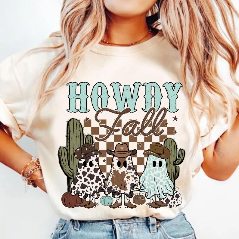 May include: White t-shirt featuring a graphic design of three ghosts wearing cowboy hats and boots. The ghosts are in front of a chequered background with the text "Howdy Fall" above them.