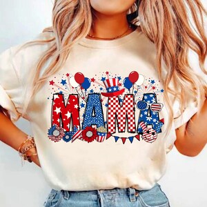 May include: A white t-shirt with a red, white, and blue graphic design that says "MAMA" with stars, fireworks, balloons, and a patriotic hat.