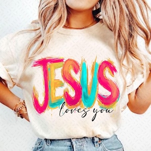 May include: Cream-colored t-shirt with the words "JESUS loves you" in a colorful, brushstroke style. The word "JESUS" is in bright pink, orange, and teal. The phrase "loves you" is in black script.