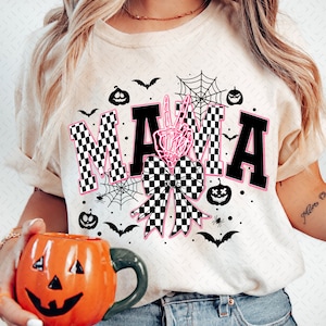 May include: A white t-shirt with a black and white checkered "MAMA" graphic. The graphic includes a pink bow with a skeleton hand peace sign in the center. The shirt is decorated with bats, spiders, and pumpkins.