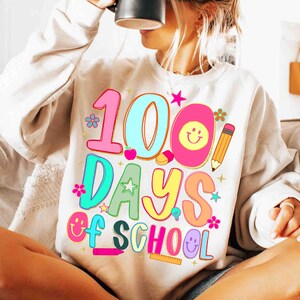 May include: A cream-coloured sweatshirt featuring a vibrant graphic that reads "100 Days of School." The design incorporates the numbers 100, the words "Days of School," and school-related illustrations such as a pencil, apple, and smiley faces. The graphic uses bright pink, blue, yellow, and green colours.