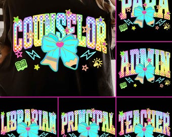 May include: Black t-shirts with colorful text and graphics. The shirts feature the words "COUNSELOR", "PARA", "ADMIN", "LIBRARIAN", "PRINCIPAL", and "TEACHER" in a checkered pattern with a bow and pencil design.