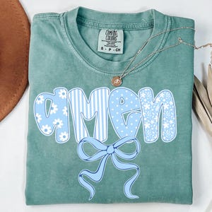May include: A seafoam green t-shirt with the word "AMEN" in large, light blue letters. The letters have different patterns, including stripes, polka dots, and floral designs. A light blue bow is below the word. A gold necklace is on top of the shirt.