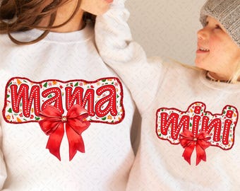 Op de afbeelding: Witte sweatshirts met rode tekst en strikken. Eén zegt "Mama" en de andere "Mini". De tekst is omlijnd met rode glitter en versierd met kerstafbeeldingen. Beide sweatshirts hebben een grote rode strik.