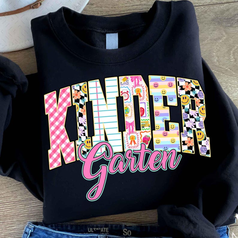 K&ouml;nnte beinhalten: Schwarzes Sweatshirt mit dem Wort "Kindergarten" in einem farbenfrohen, gemusterten Design. Das Wort "Kinder" ist in einem rosa-wei&szlig;en Vichy-Muster, floralen und Smiley-Design gehalten. Das Wort "Garten" ist in rosa Schrift geschrieben.