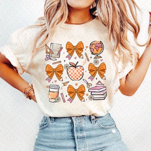 May include: A white t-shirt with a design of orange bows, a red apple, a globe, pencils, a coffee cup, scissors, a ruler, and books. The design is in a retro style with pink and yellow accents.