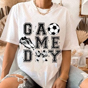 May include: White t-shirt with black glitter text that reads "GAME DAY" with a soccer ball, soccer cleats, and a bow.