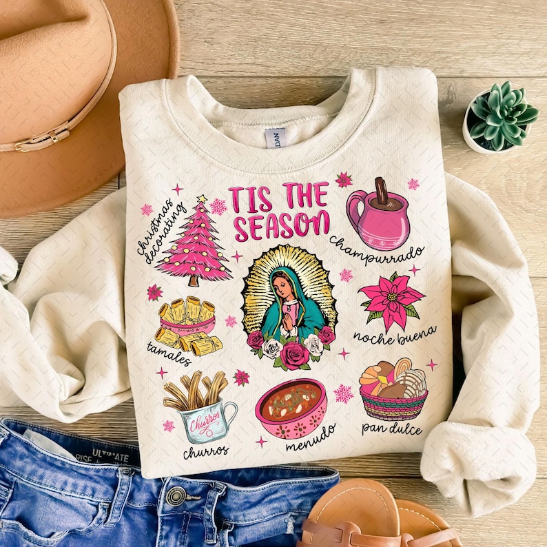May include: A beige sweatshirt with a festive Christmas design featuring a pink Christmas tree, a religious figure, and various Mexican food and drink items. The text "Tis the Season" is printed in pink letters.