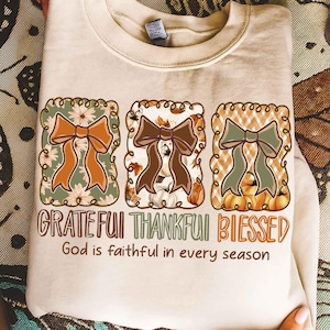 May include: Beige sweatshirt with three decorative bow designs in fall colors. The words "Grateful, Thankful, Blessed" are printed below the bows, with the phrase "God is faithful in every season" underneath.