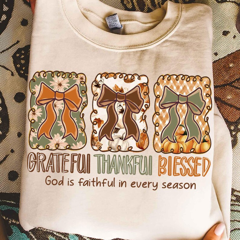 May include: Beige sweatshirt with three decorative bow designs in fall colors. The words "Grateful, Thankful, Blessed" are printed below the bows, with the phrase "God is faithful in every season" underneath.