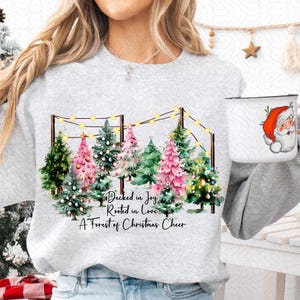 May include: A gray sweatshirt with a watercolor design of a row of Christmas trees with pink and green ornaments. The trees are decorated with string lights and the text "Decked in Joy, Rooted in Love, A Forest of Christmas Cheer" is printed below the trees.