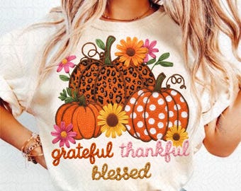 May include: Cream-colored t-shirt featuring a fall-themed design. The design includes pumpkins with leopard print and polka dots, sunflowers, and the words "grateful, thankful, blessed" in a script font. Pink and orange flowers are also included.