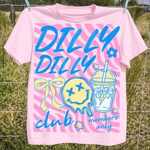 May include: Pink t-shirt with a wavy pink and white background design. The shirt features the words "DILLY DILLY CLUB" in blue script, along with a yellow melting smiley face, a bow, a drink, and the words "members only".