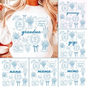 Può includere: Felpa bianca con disegni floreali blu e testo. Il design include bouquet floreali, annaffiatoi, stivali e le parole "nonna", "grammy", "gigi", "nana", "mamma" e "mimi".