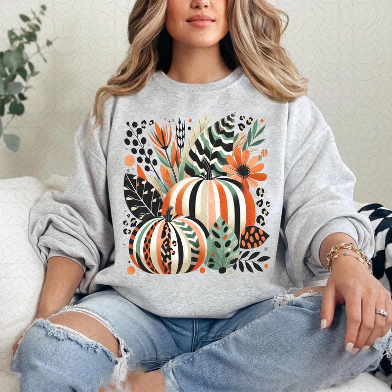 Peut inclure: Un sweat-shirt gris avec un imprim&eacute; graphique de citrouilles, de feuilles et de fleurs. Les citrouilles sont orange, blanches et noires avec un motif g&eacute;om&eacute;trique. Les feuilles sont vertes et brunes. Les fleurs sont roses et jaunes.