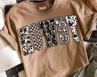 Könnte beinhalten: Ein beiges T-Shirt mit dem Wort "HOWDY" in einer kräftigen, dekorativen Schriftart. Die Buchstaben zeigen Kuhfellmuster, ein rosa-weißes Schachbrettmuster und Illustrationen zum Thema Western. Das T-Shirt ist lässig und hat kurze Ärmel.