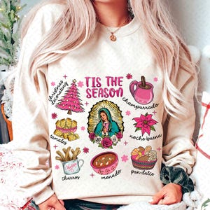 May include: A cream-colored sweatshirt with the text "Tis the Season" in pink lettering. The sweatshirt features various illustrations of traditional Mexican Christmas foods and drinks, including tamales, champurrado, churros, menudo, pan dulce, and a Christmas tree.