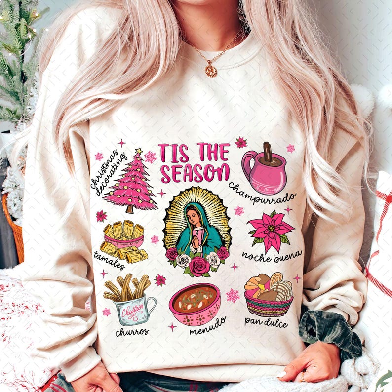 May include: A cream-colored sweatshirt with the text "Tis the Season" in pink lettering. The sweatshirt features various illustrations of traditional Mexican Christmas foods and drinks, including tamales, champurrado, churros, menudo, pan dulce, and a Christmas tree.