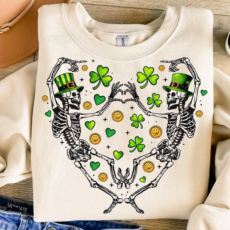 May include: A cream-colored sweatshirt with a graphic of two skeletons wearing green shamrock hats and dancing. The skeletons are surrounded by green shamrocks and gold coins.