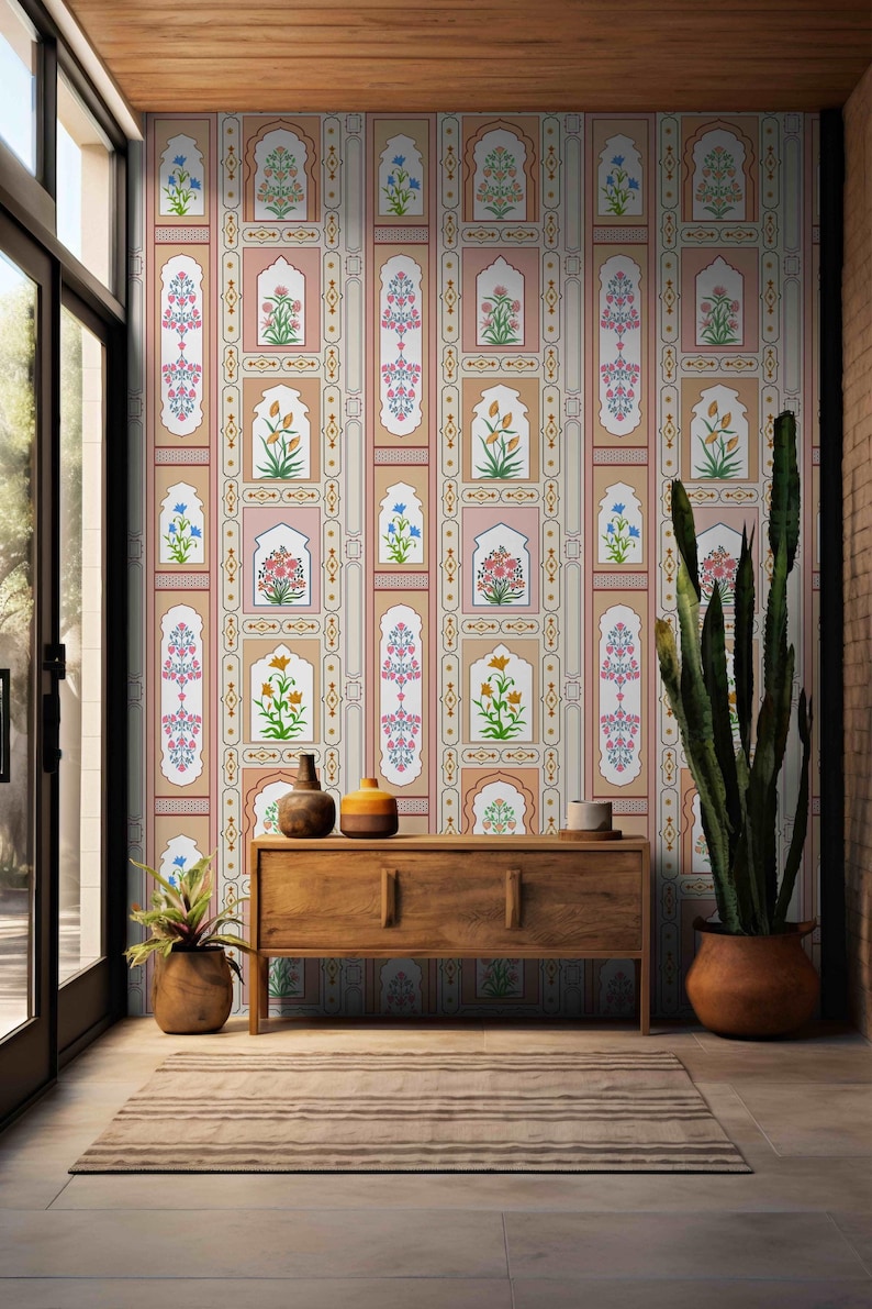 May include: A wooden console table with two drawers and a patterned wallpaper behind it. The wallpaper features a repeating design of floral motifs and arches in shades of pink, beige, and green. A potted cactus and a potted plant are on either side of the table.