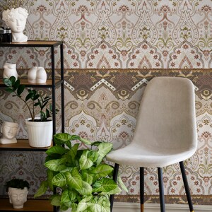 May include: A beige chair with black legs and gold accents sits in front of a wall with a brown and white patterned wallpaper. The wallpaper features a repeating design of arches and floral motifs. There are plants and decorative items on a black metal shelf in the foreground.