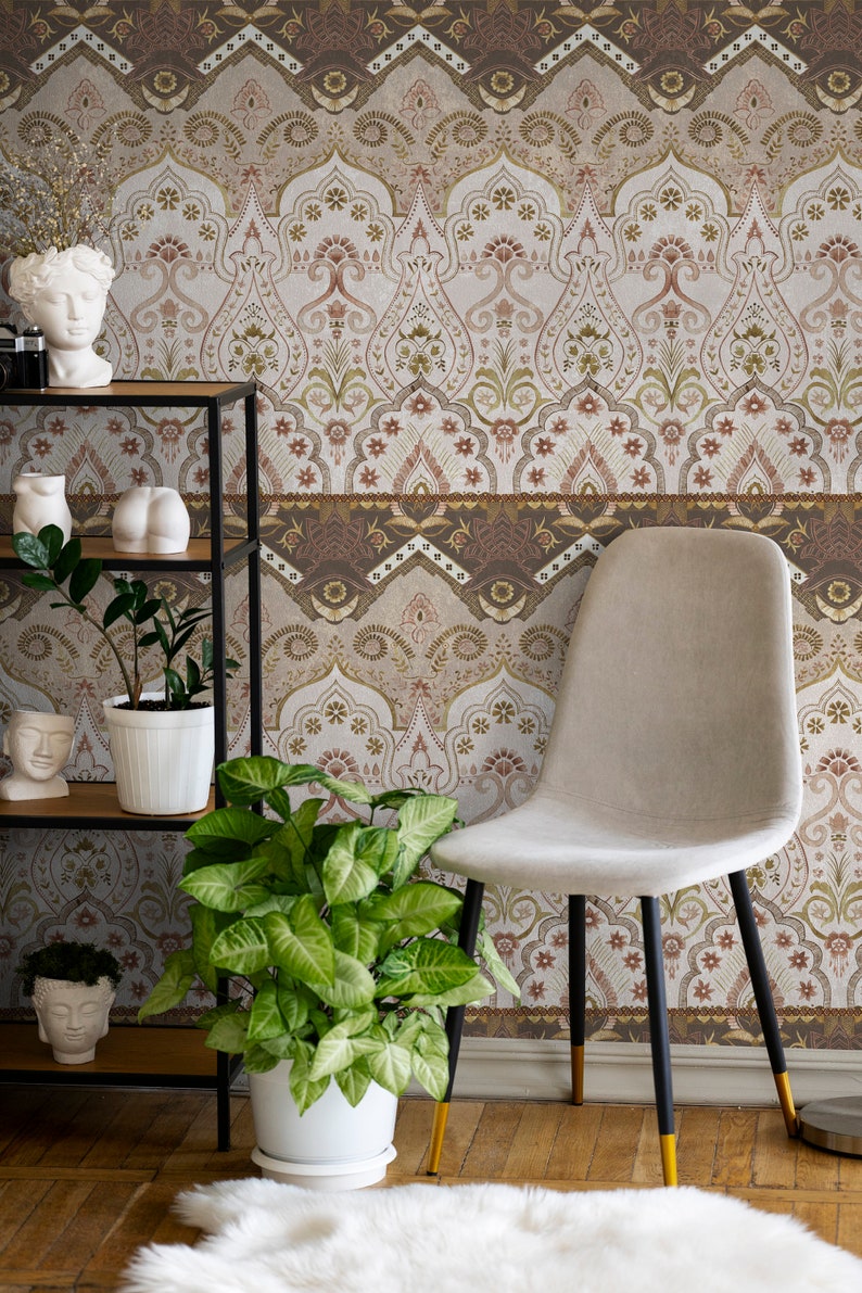 May include: A beige chair with black legs and gold accents sits in front of a wall with a brown and white patterned wallpaper. The wallpaper features a repeating design of arches and floral motifs. There are plants and decorative items on a black metal shelf in the foreground.