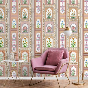 May include: A pink armchair with a gold metal frame sits in front of a wall with a patterned wallpaper. The wallpaper features a repeating design of arches with floral motifs in shades of pink, orange, yellow, blue, and green. A floor lamp with a gold metal base and a white globe shade stands to the right of the chair.