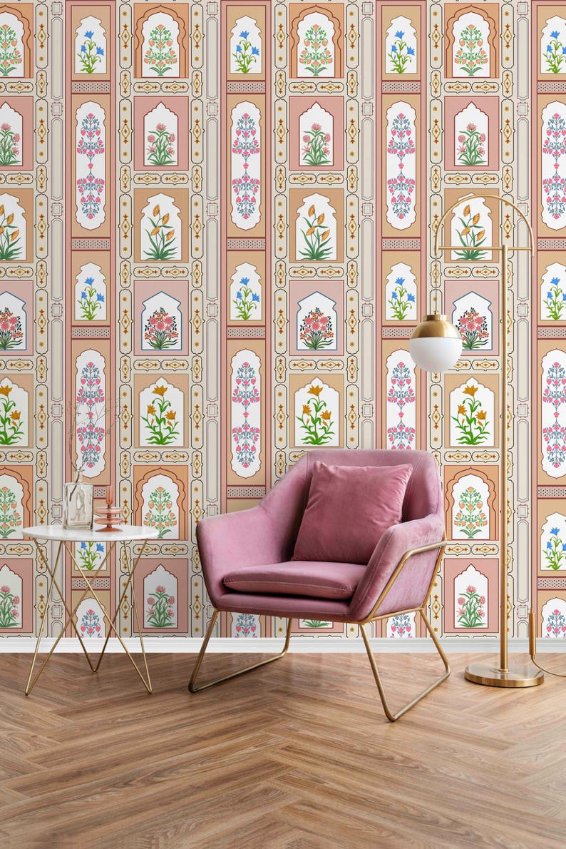 May include: A pink armchair with a gold metal frame sits in front of a wall with a patterned wallpaper. The wallpaper features a repeating design of arches with floral motifs in shades of pink, orange, yellow, blue, and green. A floor lamp with a gold metal base and a white globe shade stands to the right of the chair.