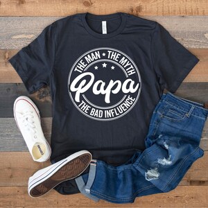 May include: A black t-shirt with a white circle graphic that says "The Man * The Myth * Papa * The Bad Influence".