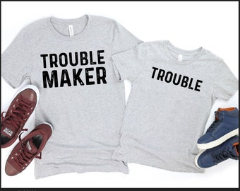 May include: Matching gray t-shirts with the text "Trouble Maker" on the larger shirt and "Trouble" on the smaller shirt. The shirts are laid out on a white background with a pair of brown sneakers and a pair of blue sneakers.
